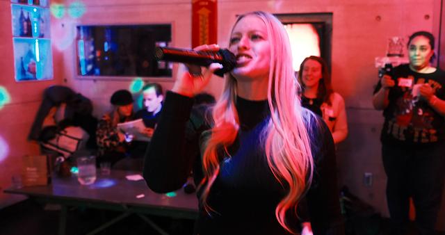 Blonde host singing into the mic with guests gathered close behind her