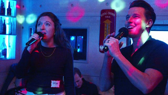 A guest and a Broadway host trading verses on a duet, each on a mic under blue and red light in the intimate room at RPM Underground
